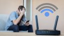 EIn Mann schaut frustriert auf sein handy während der Router geringe Internetgeschwindigkeit anzeigt.