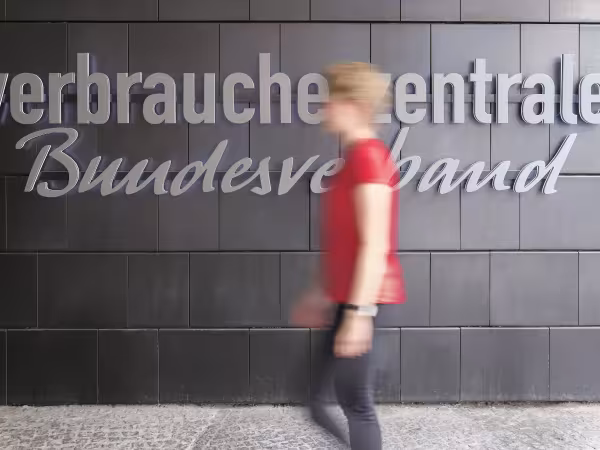 Zu sehen ist eine blonde Frau, bekleidet mit einem roten T-Shirt und einer schwarzen Hose, die vor einer schwarzen Wand langläuft. Die Frau ist durch die Bewegung unscharf. Auf der schwarzen Wand ist der graue Schriftzug "Verbraucherentrale Bundesverband" zu lesen ist. Es handelt sich um den Eingangsbereich des vzbv-Gebäudes.