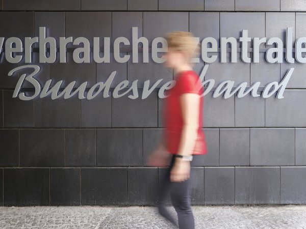 Zu sehen ist eine blonde Frau, bekleidet mit einem roten T-Shirt und einer schwarzen Hose, die vor einer schwarzen Wand langläuft. Die Frau ist durch die Bewegung unscharf. Auf der schwarzen Wand ist der graue Schriftzug "Verbraucherentrale Bundesverband" zu lesen ist. Es handelt sich um den Eingangsbereich des vzbv-Gebäudes.