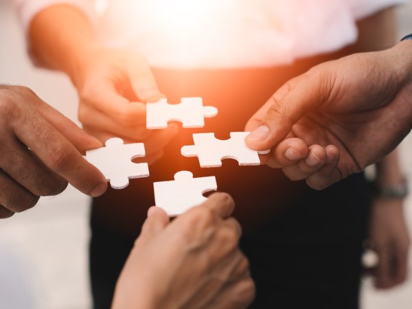 Zu sehen ist jeweils die rechte Hand von vier unterschiedlichen, stehenden Personen mit einem weißen Puzzleteil. Die Hände der Personen nähern sich in der Mitte an, um die Puzzleteile zu einem Ganzen zusammenzufügen.
