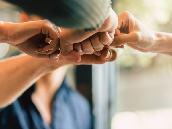 Zu sehen ist, wie eine Gruppe von Menschen unterschiedlichen Alters, Geschlecht und Herkunft die lockere Fäuste einer Hand in ihrer Mitte zusammenstoßen. Teilweise sind die Ärmel der Kleidungsstücke zu erkennen. Der Hintergrund ist unscharf.