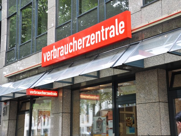Zu sehen ist das Schild der Verbraucherzentrale an einer Gebäudefassade über dem Eingangsbereich. Es handelt sich um das Gebäude der Verbraucherzentrale NRW. Auf rotem Grund ist mit weißen Buchstaben das Wort "Verbraucherzentrale" zu lesen. 