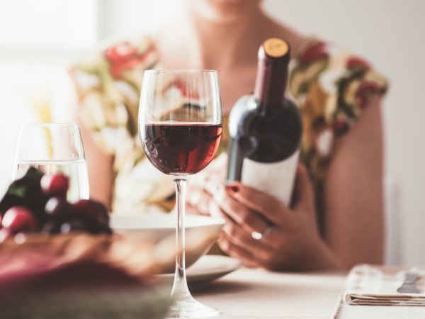 Frau betrachtet Weinflasche in ihrer Hand, im Vordergrund ein Glas Rotwein