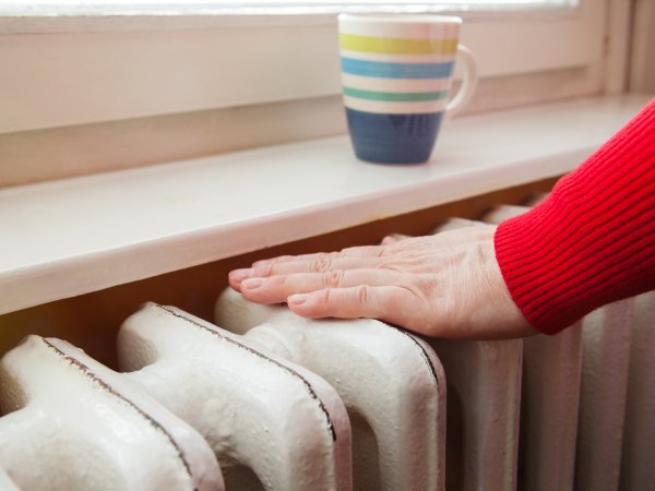 Zu sehen ist eine Heizung unter einer Fensterbank. Auf der Fensterbank steht ein Kaffeebecher. Eine Person wärmt sich die Hände auf der Heizung. Von der Person ist nur eine Hand und ein Unterarm abgebildet. 