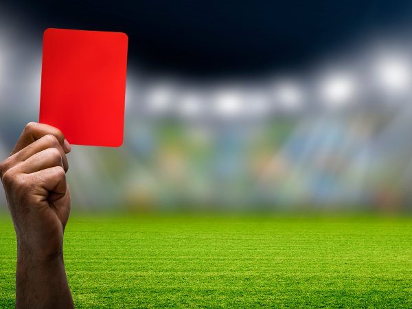 Eine Hand hält eine rote Karte hoch. Im Hintergrund sieht man ein Fußballstadion.