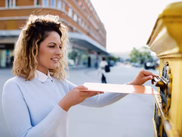 Zu sehen ist eine junge Frau, die einen Umschlag in einen gelben Briefkasten wirft. Der Briefkasten ist nur im Anschnitt zu sehen. Im Hintergrund der lächelnden Frau sind Gebäude einer Stadt zu erkennen. 