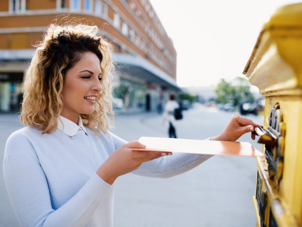 Zu sehen ist eine junge Frau, die einen Umschlag in einen gelben Briefkasten wirft. Der Briefkasten ist nur im Anschnitt zu sehen. Im Hintergrund der lächelnden Frau sind Gebäude einer Stadt zu erkennen. 