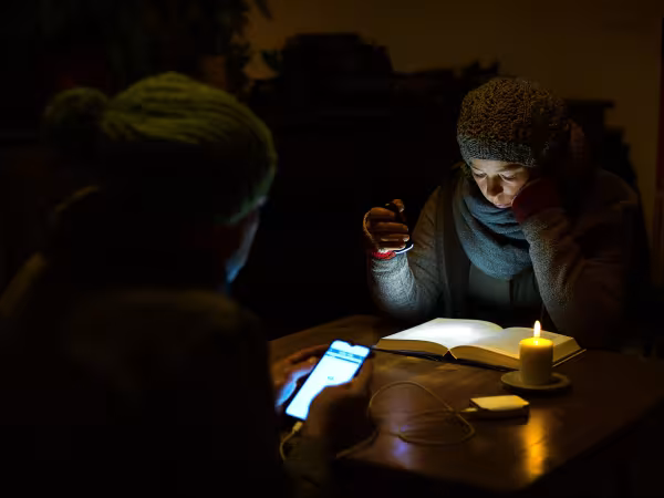 Ein Mann und eine Frau sitzen dick angezogen in einem dunklen Wohnzimmer bei Kerzenschein. Sie liest mit einer Taschenlampe ein Buch. Er sieht auf sein Handy