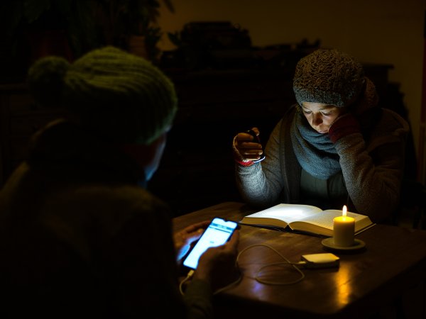 Ein Mann und eine Frau sitzen dick angezogen in einem dunklen Wohnzimmer bei Kerzenschein. Sie liest mit einer Taschenlampe ein Buch. Er sieht auf sein Handy