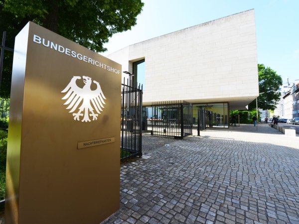 Am linken Bildrand ist eine Stele abgebildet, auf der oben Bundesgerichtshof steht. Darunter ist der Bundesadler zu sehen. Unterhalb des Bundesadlers befindet sich der Nachtbriefkasten des BGH. Im rechten Bildausschnitt ist eine Auffahrt sowie ein Gebäude im  Hintergrund zu sehen. 