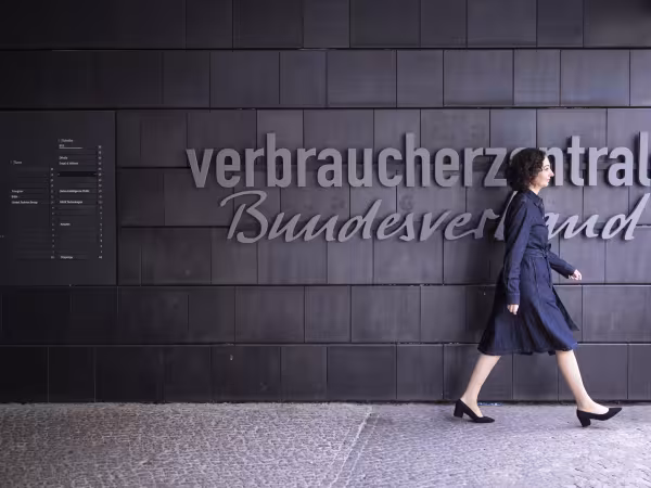 Zu sehen ist Ramona Pop, die vor einer dunklen Wand entlangläuft. Auf der Wand steht "Verbraucherzentrale Bundesverband". Es handelt sich um den Eingang des vzbv-Gebäudes. 