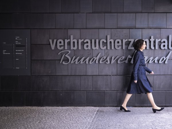 Zu sehen ist Ramona Pop, die vor einer dunklen Wand entlangläuft. Auf der Wand steht "Verbraucherzentrale Bundesverband". Es handelt sich um den Eingang des vzbv-Gebäudes. 