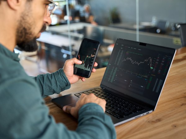 Ein Mann sitzt am Tisch, hält ein Handy in der Hand und blickt auf Aktienverläufe auf dem Handy und dem Laptop, der vor ihm steht.