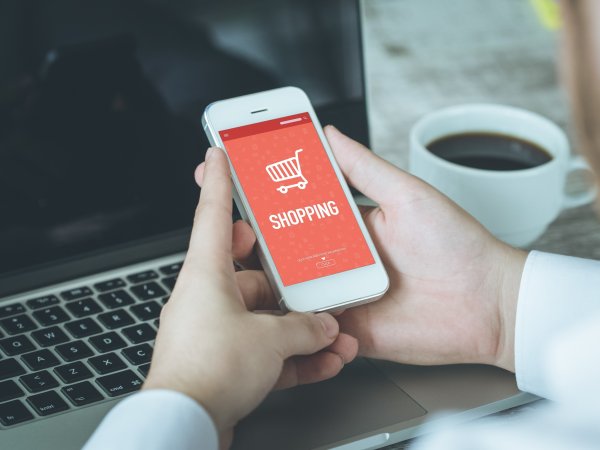 Smartphone in der Hand vor Laptop, Onlineshop auf Handydisplay in weißer Schrift auf orangen Hintergrund