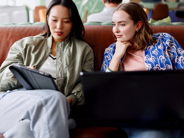 Zwei Frauen sitzen auf einem Sofa; die eine Frau hat einen Laptop auf dem Schoß, die andere hat ein Tablet in der Hand.
