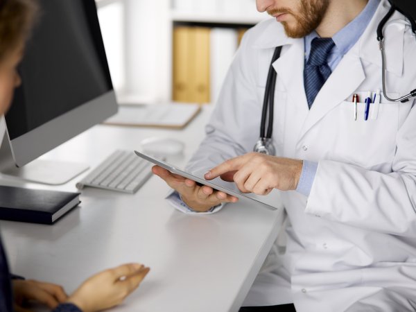 Arzt und Patientin sitzen am Tisch; der Arzt schaut auf ein Tablet
