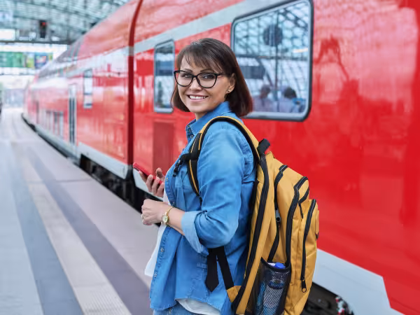 Frau mit Handy in der Hand dreht sich lächend nach hinten auf dem Bahnsteig, rechts neben ihr steht ein Zug der Deutschen Bahn