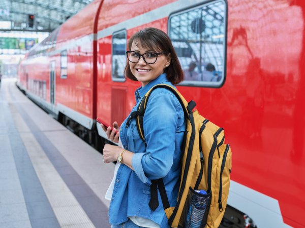 Frau mit Handy in der Hand dreht sich lächend nach hinten auf dem Bahnsteig, rechts neben ihr steht ein Zug der Deutschen Bahn