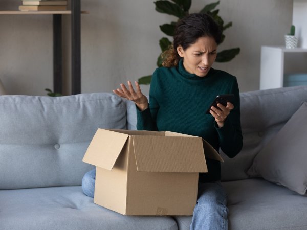 Frau sitzt auf Sofa und hat ein Paket vor sich stehen; sie blickt ärgerlich auf ihr Handy, das sie in der Hand hält