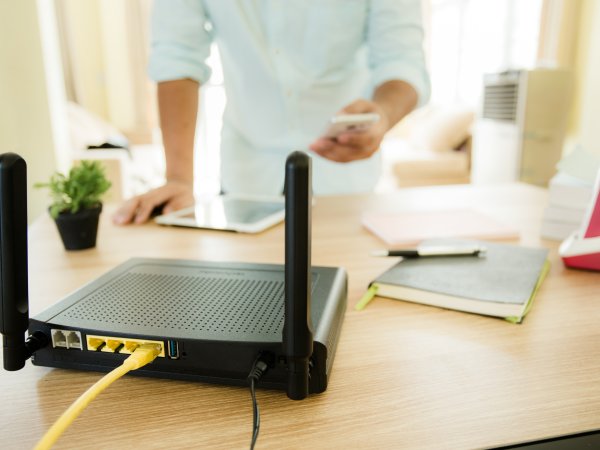 Auf dem Tisch steht im Vorderund ein Router, im Hintergrund ist eine Person mit einem Handy in der Hand zu erkennen