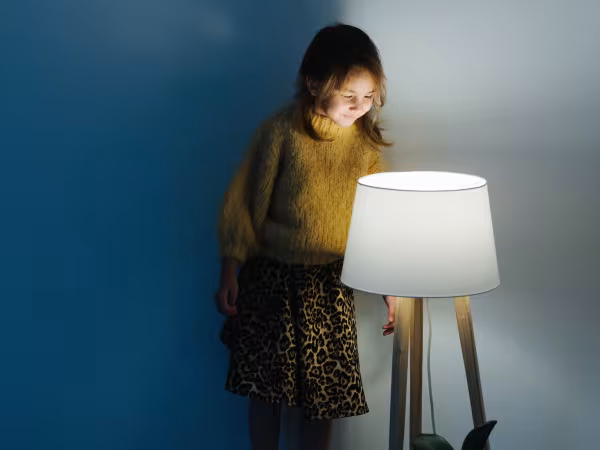 Ein kleines Mädchen steht auf der Sofakante und schaut von oben in die brennende Stehlampe. 