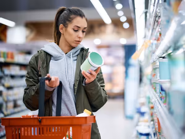 Eine Frau steht im Supermarkt vorm Regal und blickt auf eine Dose, die sie in der Hand hält