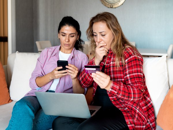 Zwei Frauen sitzen auf dem Sofa. Eine von beiden hat einen Laptop vor sich und eine Kreditkarte in der Hand. Die andere hält ein Handy in der Hand. Beide schauen bestürzt.