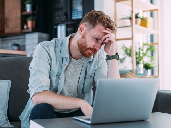 Ein Mann sitzt über einen Laptop gebeugt am Tisch und hält sich die Hand vor der Stirn verschränkt