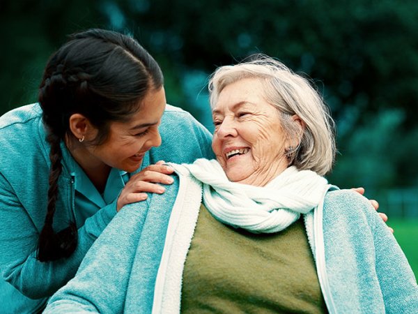 Junge Pflegerin beugt sich zu älterer im Rollstuhl sitzender Dame hinunter. 