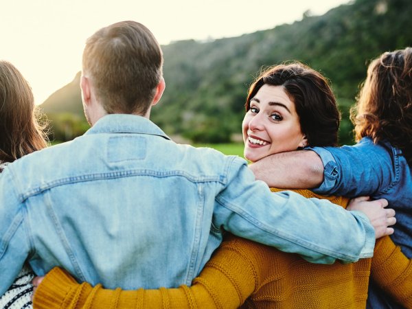 4 Menschen stehen sich in den Armen haltend zusammen; ihre Blicke sind nach vorne (für den:die Betrachter:in nicht sichtbar) abgewandt; eine Person dreht sich zum:zur Betrachter:in um