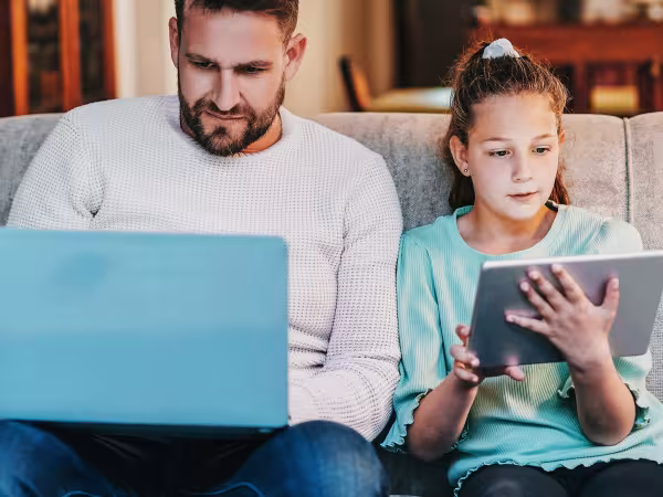 Ein Mann und ein Kind sitzen nebeneinander auf dem Sofa. Der Mann hat einen Laptop auf dem Schoß, das Mädchen ein Tablet in der Hand.