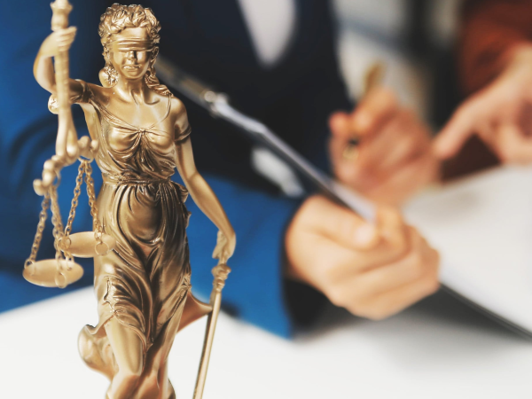 Eine Justitia-Figur steht auf dem Tisch; dahinter arbeiten zwei Personen an Dokumenten.