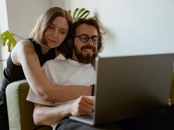 Mann sitzt auf dem Sofa vor dem Laptop; Frau umarmt ihn von hinten und schaut ebenfalls auf den Laptop.
