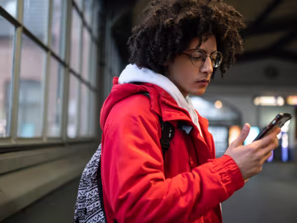 Ein Jugendlicher steht in einer U-Bahn-Station und blickt auf sein Handy, das er in der Hand hält.