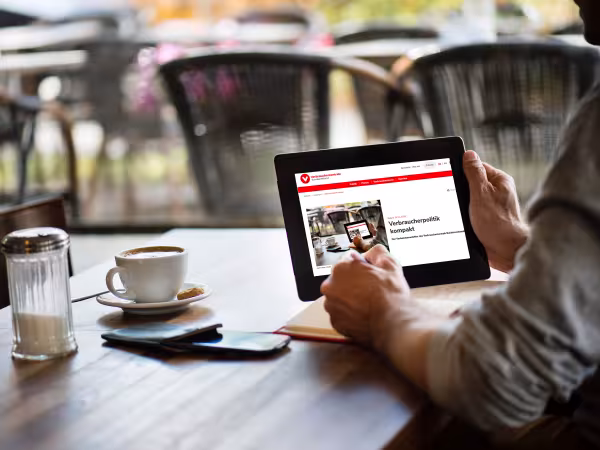 Ein Mann sitzt an einem Tisch, trinkt einen Kaffee und liest den Newsletter Verbraucherpolitik kompakt auf einem Tablet