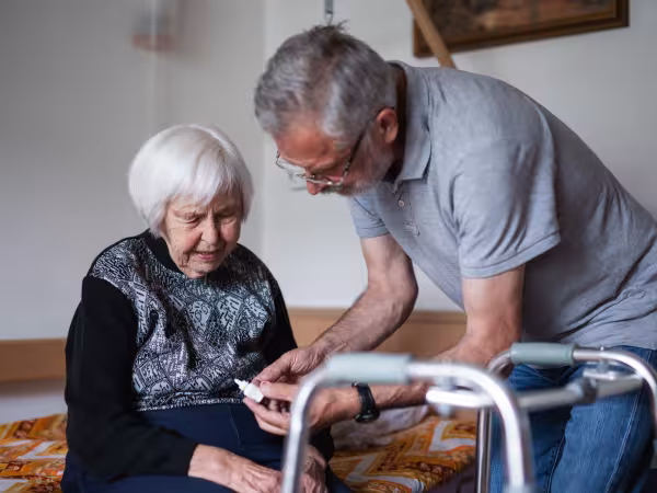 Eine Seniorin sitzt auf einem Pflegebett. Vor ihr steht ein Mann, der ihr ein Medikament gibt. Vor der Frau steht eine Gehhilfe.