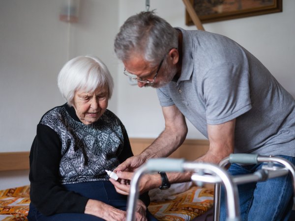 Eine Seniorin sitzt auf einem Pflegebett. Vor ihr steht ein Mann, der ihr ein Medikament gibt. Vor der Frau steht eine Gehhilfe.
