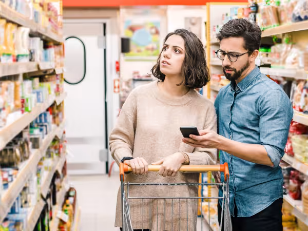 Ein Paar steht im Supermarkt mit Einkaufswagen. Die Frau schaut zum Regal, während der Mann in sein Handy guckt.