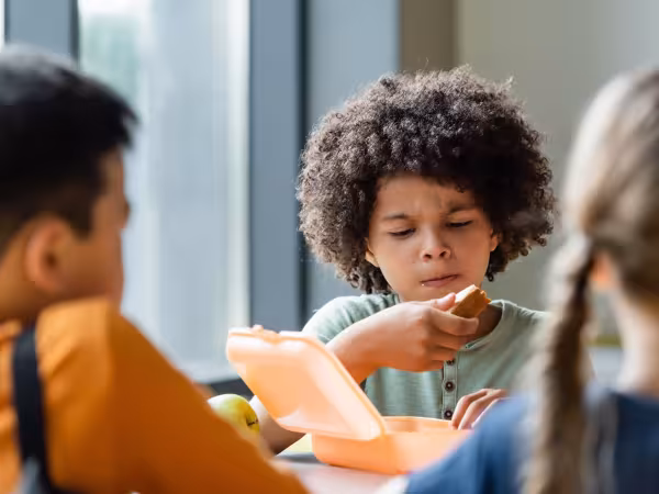Junge isst an Tisch mit zwei weiteren Kindern Pausenbrot