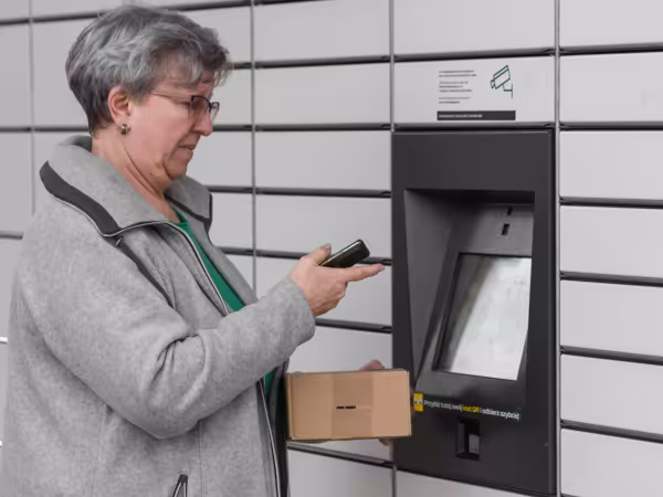 Ältere Frau steht vor einer Packstation. In den Händen hält sie ein Handy und ein Paket.