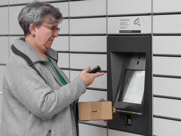 Ältere Frau steht vor einer Packstation. In den Händen hält sie ein Handy und ein Paket.