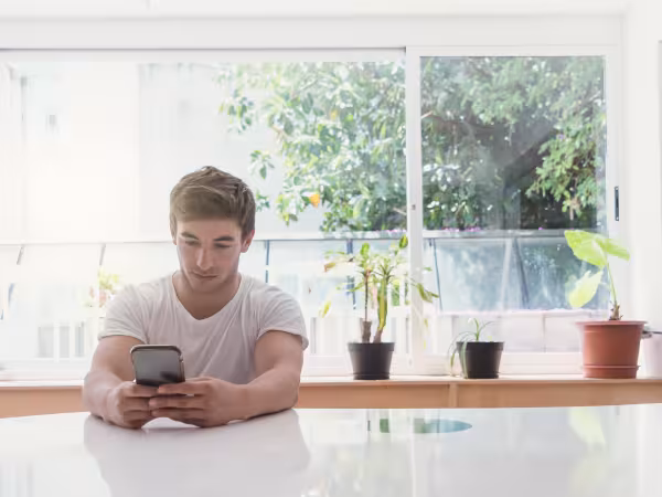 junger Mann sitzt in Küche und schaut auf sein Handy