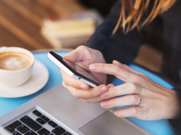 Person hält Handy vor Laptop, daneben steht ein Cappuccino 