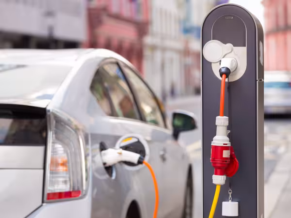 Zu sehen ist ein parkendes Auto, das an einer Ladesäule angeschlossen ist. Das silberne Auto ist nur von hinten und teilweise sichtbar, da es auch unscharf ist. Die Ladesäule mit dem orange-roten Kabel ist hingegen sehr prägnant und klar. 