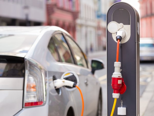 Zu sehen ist ein parkendes Auto, das an einer Ladesäule angeschlossen ist. Das silberne Auto ist nur von hinten und teilweise sichtbar, da es auch unscharf ist. Die Ladesäule mit dem orange-roten Kabel ist hingegen sehr prägnant und klar. 
