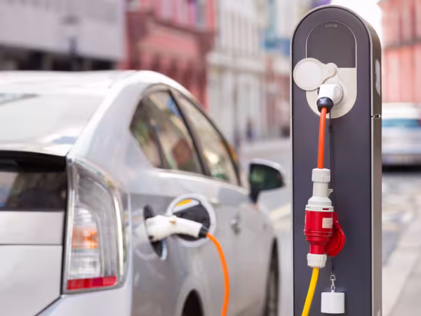 Zu sehen ist ein parkendes Auto, das an einer Ladesäule angeschlossen ist. Das silberne Auto ist nur von hinten und teilweise sichtbar, da es auch unscharf ist. Die Ladesäule mit dem orange-roten Kabel ist hingegen sehr prägnant und klar. 