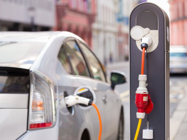 Zu sehen ist ein parkendes Auto, das an einer Ladesäule angeschlossen ist. Das silberne Auto ist nur von hinten und teilweise sichtbar, da es auch unscharf ist. Die Ladesäule mit dem orange-roten Kabel ist hingegen sehr prägnant und klar. 
