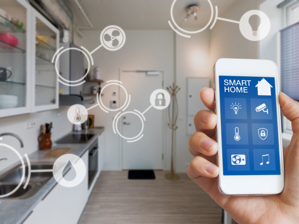 Eine Hand hält ein Handy, auf dem "Smart Home" steht. Dahinter sieht man eine Küche, in der verschiedene Smart-Home-Geräte markiert sind.