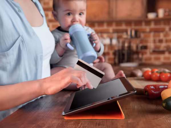 Baby sitz auf einem Tisch, festgehalten von eine Person und trinkt aus einer Flasche; Die Person auf Tablet und hält mit der rechten Hand eine Bankkarte