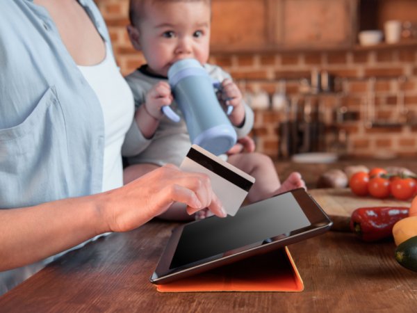 Baby sitz auf einem Tisch, festgehalten von eine Person und trinkt aus einer Flasche; Die Person auf Tablet und hält mit der rechten Hand eine Bankkarte